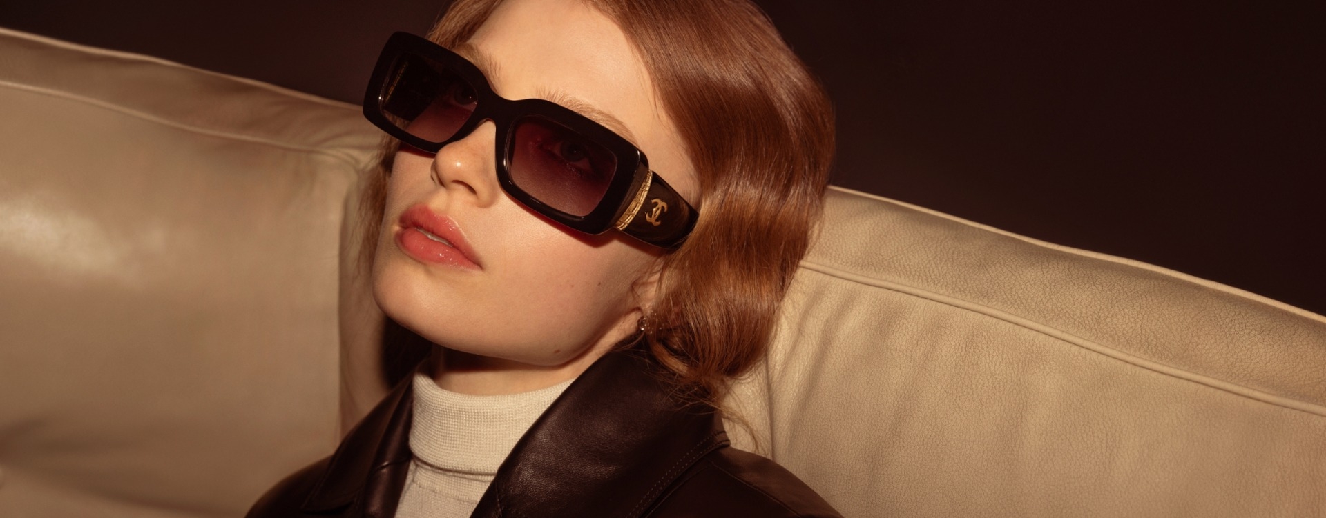 Frau trägt elegante, große Sonnenbrille mit goldenem Chanel-Logo und sitzt auf einem cremefarbenen Ledersofa.