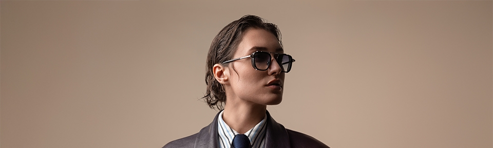 Eine stilvolle Person mit zurückgekämmtem, nassem Haar trägt eine elegante John Dalia Sonnenbrille und ein formelles Outfit bestehend aus Hemd, Krawatte und Sakko. Der Hintergrund ist neutral beige.