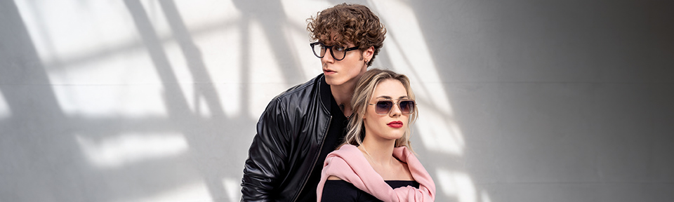 Stylisches Paar mit Brillenmode: er in Lederjacke, sie mit Sonnenbrille und rosa Pullover über den Schultern.
